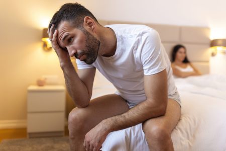 Young couple having a problem. Guy is sitting on bed and looking sadly away, his girlfriend in the background. Upset young couple having problems with sex